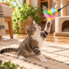 Kucing kampung tabby abu-abu kecokelatan dengan dada dan kaki putih sedang bermain dengan mainan tongkat bulu warna-warni di atas karpet krem. Kucing mengangkat satu kaki depan untuk meraih bulu berwarna pink, kuning, hijau, dan biru yang dilengkapi lonceng kecil. Suasana ruang keluarga estetik dengan cahaya matahari alami, tanaman hijau, furnitur kayu, dan cat tree di latar belakang.